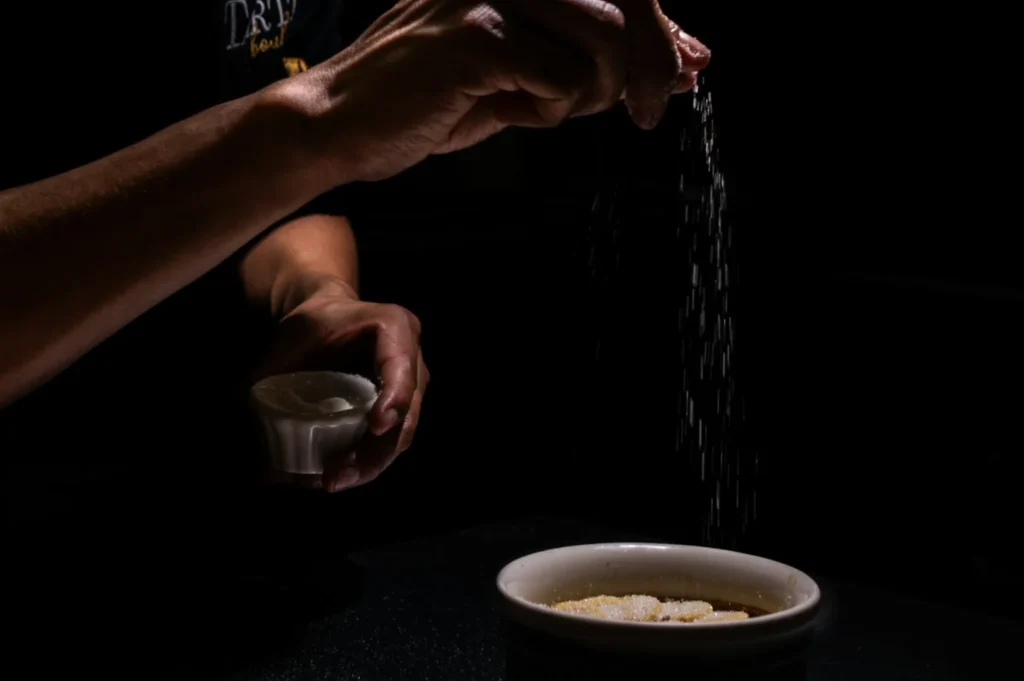 Hands sprinkle sugar over a dish in low lighting, contrasting shadows create a focused and intimate culinary scene, evoking precision and care.