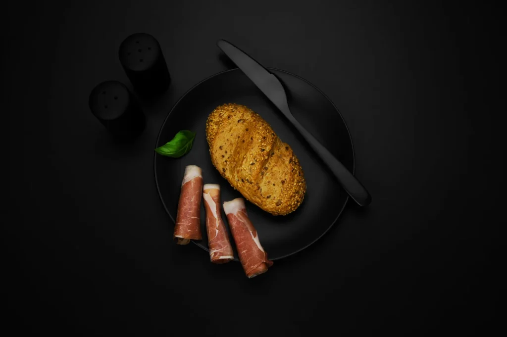 A black plate on a dark surface holds seeded bread, a basil leaf, and rolled cured meat. A knife is on the plate, and salt and pepper shakers are nearby.