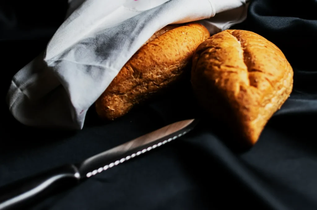 Two rustic loaves of bread partially covered with a white cloth sit on a dark surface. A serrated knife lies nearby, creating a cozy, inviting scene.