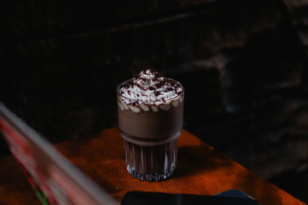A glass of creamy chocolate milkshake topped with whipped cream and chocolate shavings sits on a wooden table, evoking a cozy, indulgent mood.
