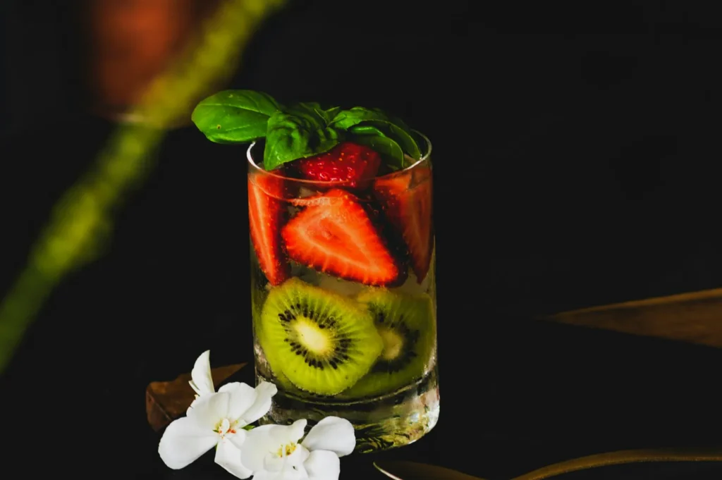 A refreshing glass filled with sliced strawberries, kiwis, and basil leaves on a dark background, accompanied by white flowers, conveying a fresh, vibrant tone.
