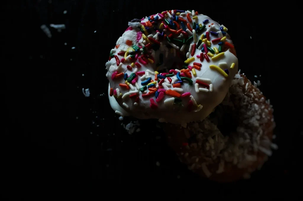 Two donuts on a dark surface; one covered in white icing with colorful sprinkles, the other with shredded coconut, creating a tempting contrast.