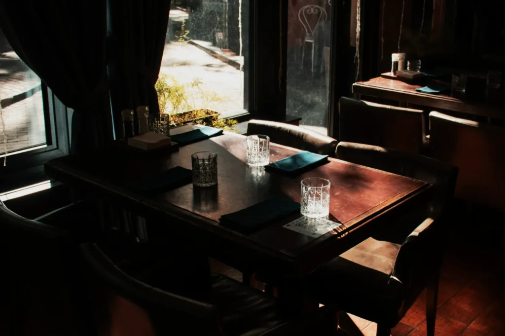 Cozy restaurant interior with wooden tables set for four. Soft sunlight filters through large windows, casting warm shadows. Dark blue napkins and crystal glasses are arranged neatly, creating an inviting and intimate ambiance.