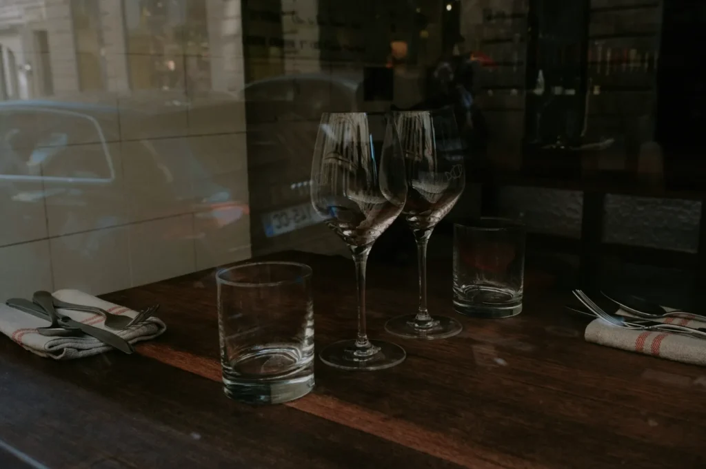 A dimly lit restaurant table set with two empty wine glasses, two empty tumblers, and neatly folded napkins with cutlery, viewed through a window.