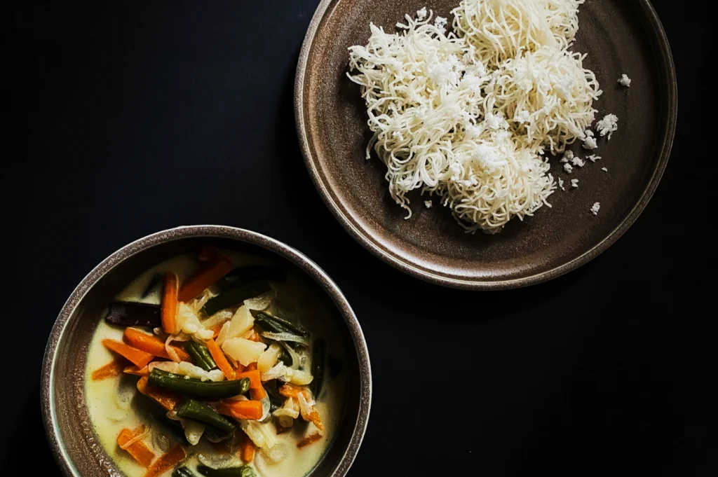 Two brown bowls on a black surface: one with string hoppers and coconut, the other with vegetable curry in a creamy yellow sauce, creating a cozy, inviting scene.