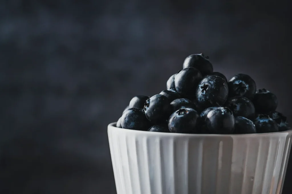 A white ceramic bowl filled with fresh blueberries against a dark, blurred background. The setting conveys a fresh and serene mood.