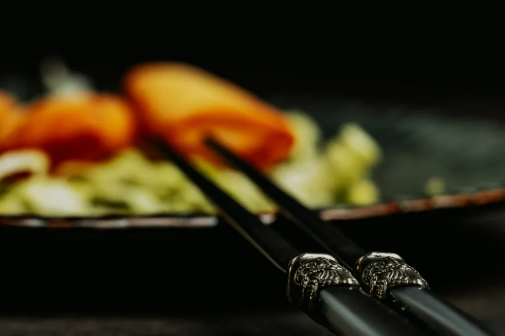 Close-up of decorative black chopsticks resting on a plate with blurred vegetables, including orange and green hues, creating a serene, elegant dining scene.