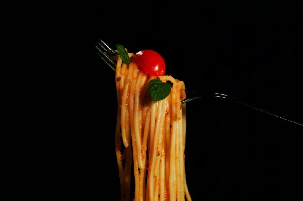 A fork holds a swirl of spaghetti in tomato sauce, topped with a cherry tomato and basil leaf, set against a black background.