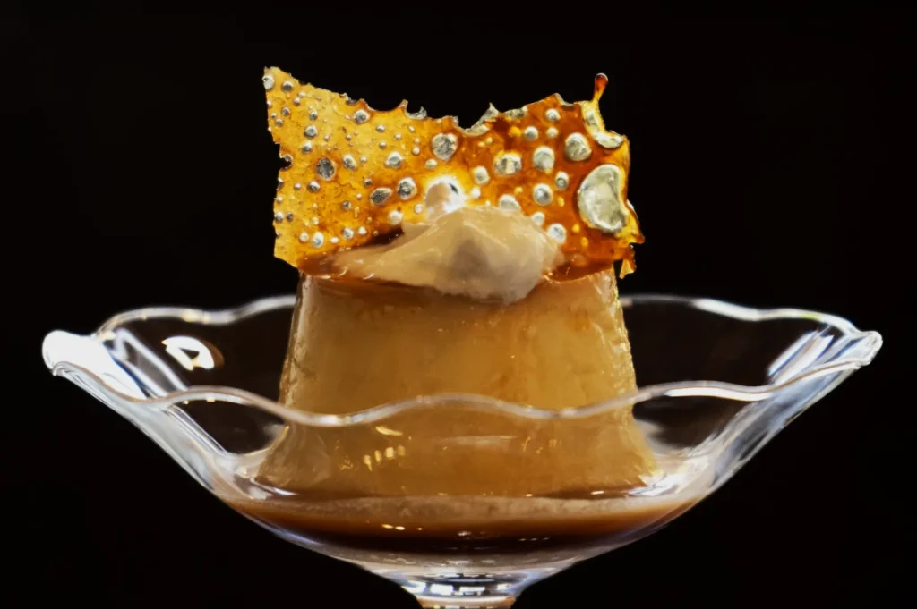 Elegant caramel flan in a glass dish, topped with a crispy, golden sugar shard and creamy dollop, set against a dark background. Decadent and refined.