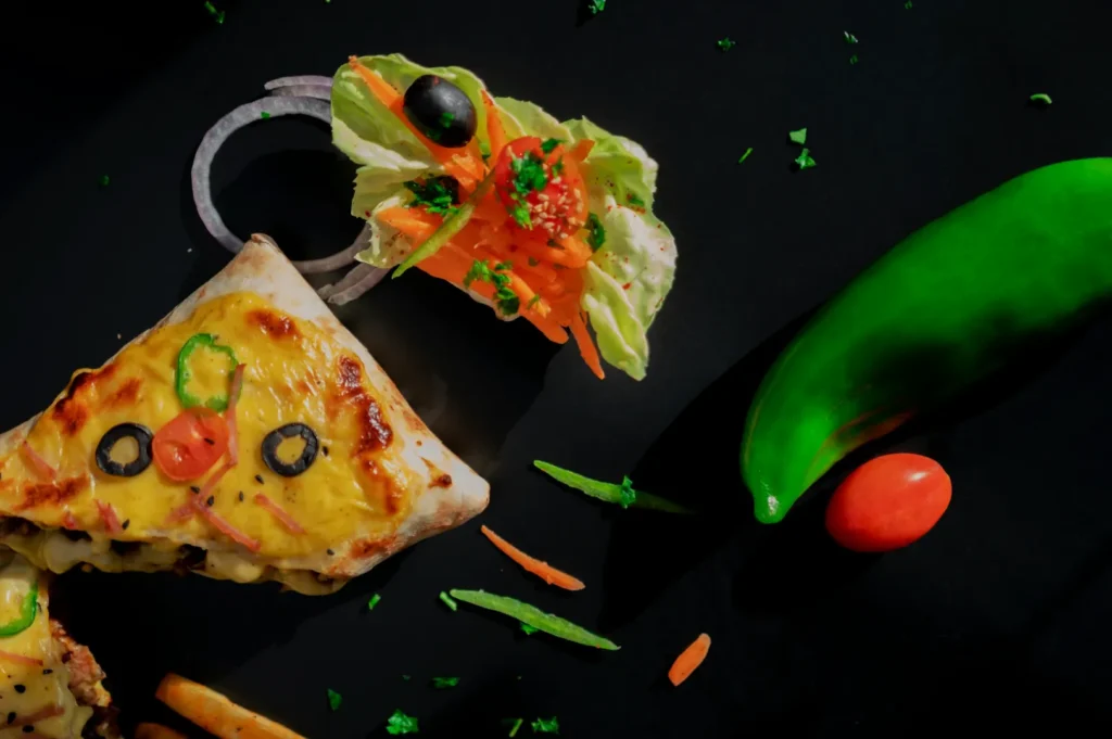 Creative pizza slice designed as an owl face with cheese, bell peppers, and olives. Beside it are a lettuce garnish, a green chili, and a small tomato.