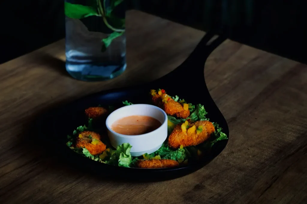 Black plate with crispy fried food on a bed of lettuce surrounding a white bowl of dipping sauce. A glass with water and leaves is in the background.
