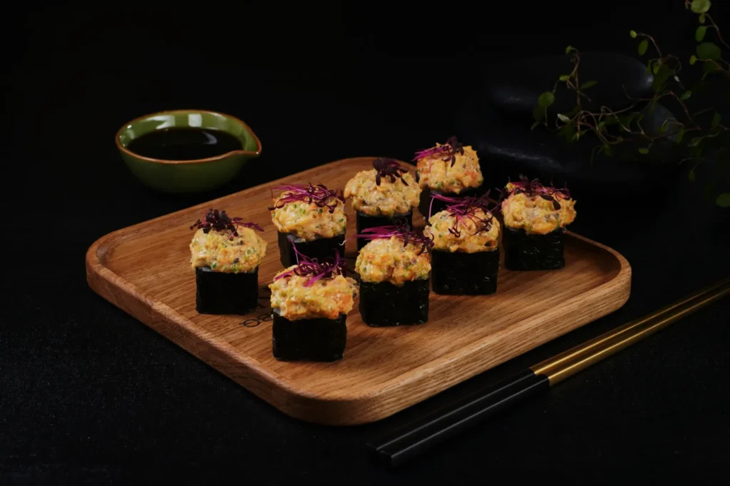 A wooden tray with eight sushi pieces wrapped in seaweed, topped with a creamy filling and purple sprouts. A green sauce dish and chopsticks on the side.