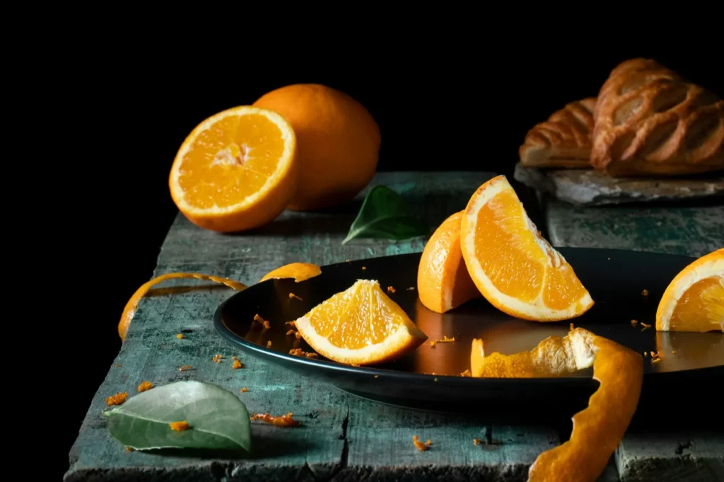 Slices of juicy orange arranged on a dark platter with orange peel and green leaves, set on a rustic wooden table. A croissant rests in the background.