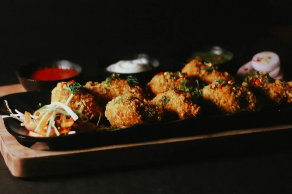 Close-up of a black tray with crispy golden fritters garnished with herbs. Side of sliced onions and dipping sauces, set on a wooden board.