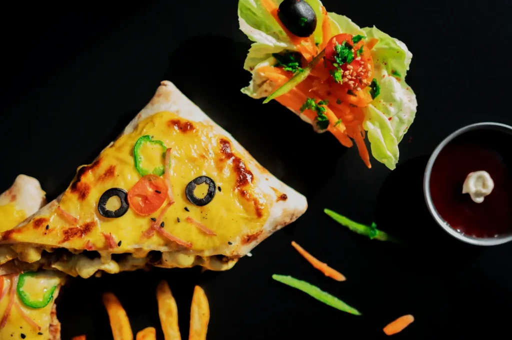 A playful food arrangement showing pizza slices with cheese, olives, and peppers resembling faces, paired with fries, salad, and a dipping sauce.