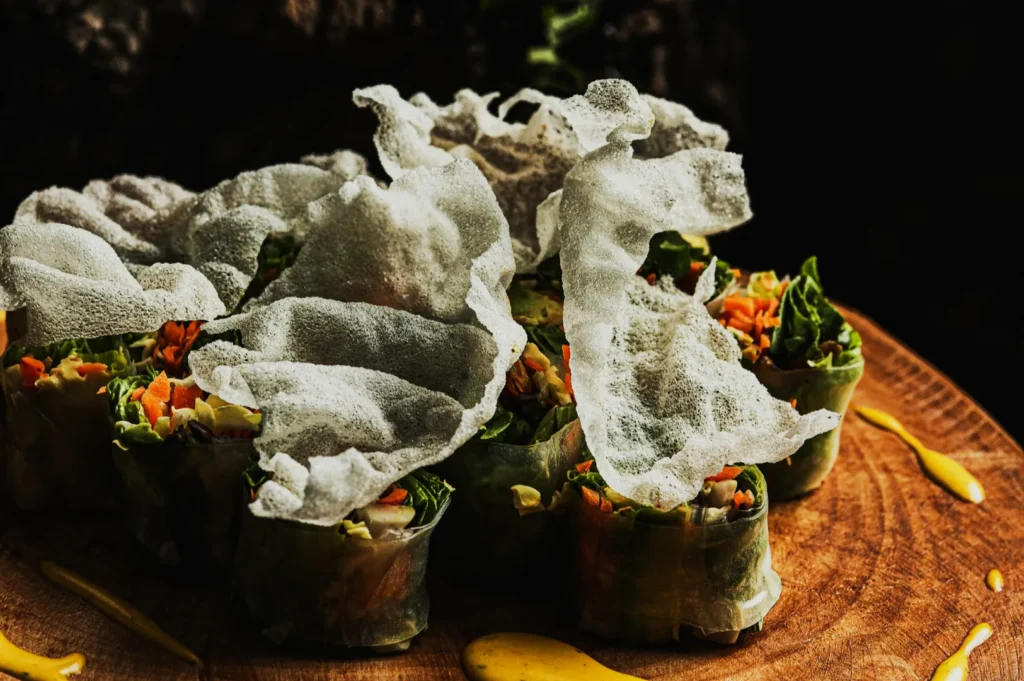 Vegetable spring rolls on a wooden board, topped with delicate, crispy rice paper. Yellow sauce drizzle adds vibrant contrast. Inviting and fresh.