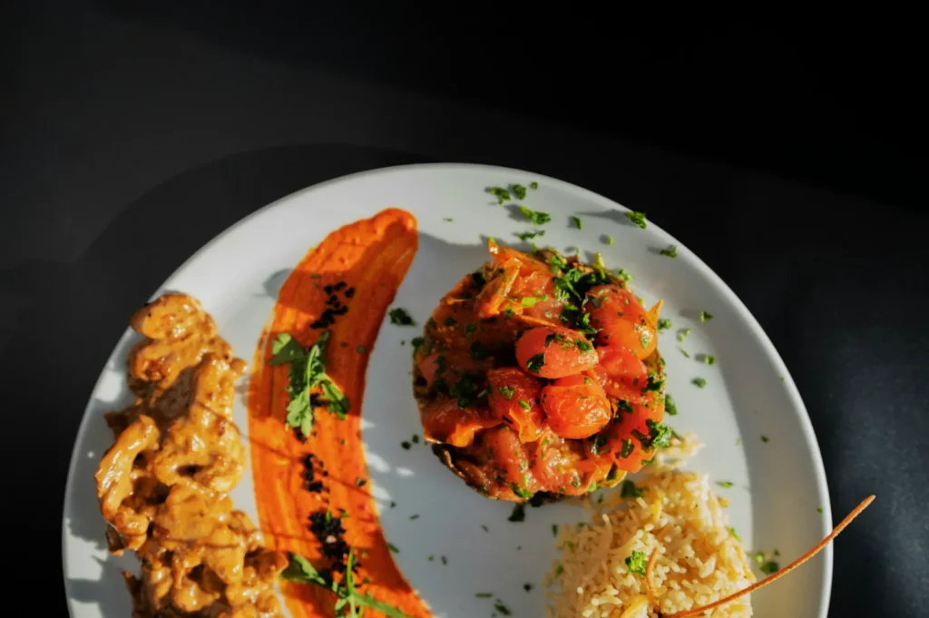 A beautifully plated dish with creamy beef, vibrant red sauce with herbs, a stack of seasoned tomatoes, and a side of fluffy rice on a white plate.