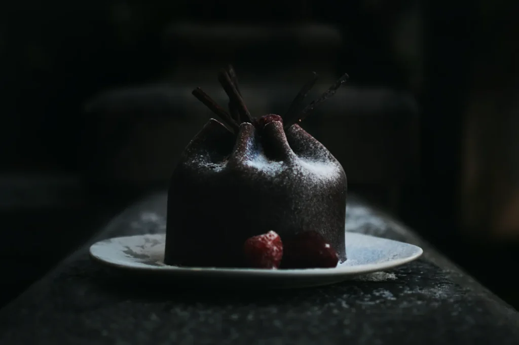A rich, chocolate dessert shaped like a dome rests on a white plate. It is topped with cocoa sticks and powdered sugar, accompanied by red berries. The dark, moody setting enhances its elegance.