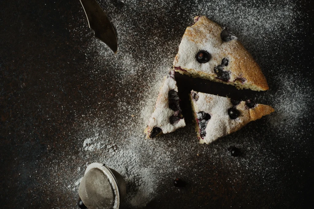 Overhead view of two blueberry cake slices dusted with powdered sugar on a dark surface, alongside a vintage knife and tea strainer. Moody ambiance.
