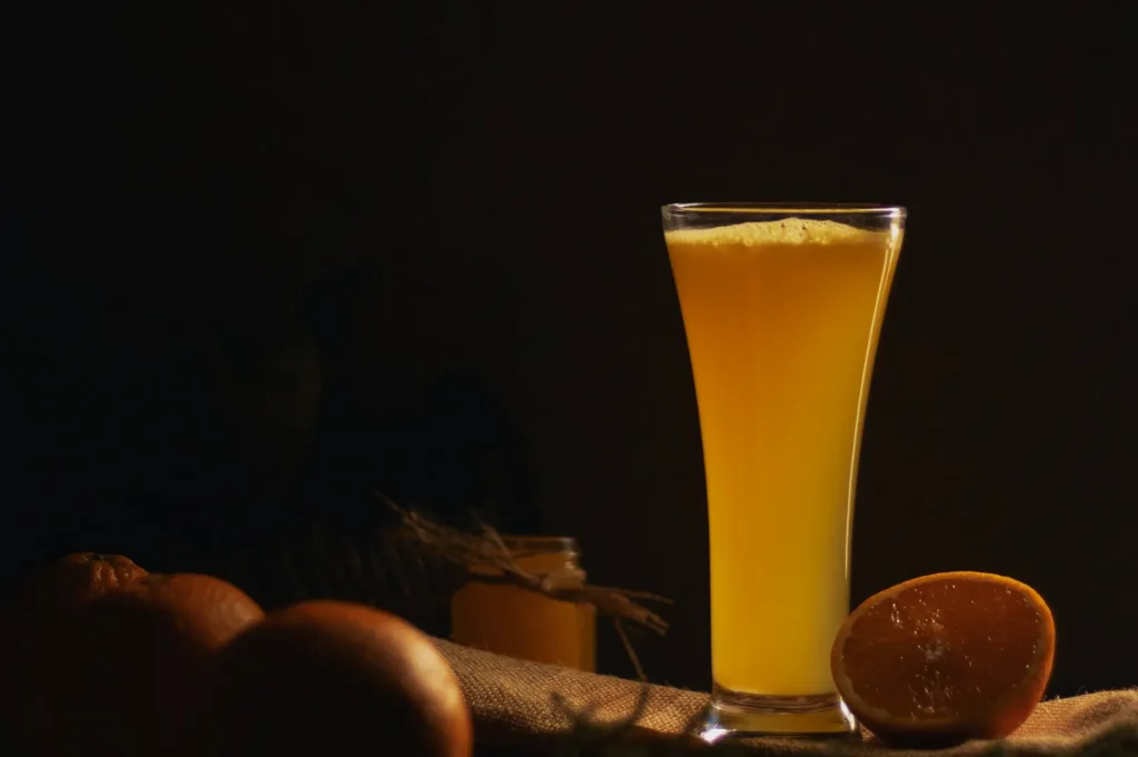 A glass of vibrant orange juice sits on a rustic cloth, with whole and halved oranges nearby. The dim lighting creates a warm, inviting atmosphere.