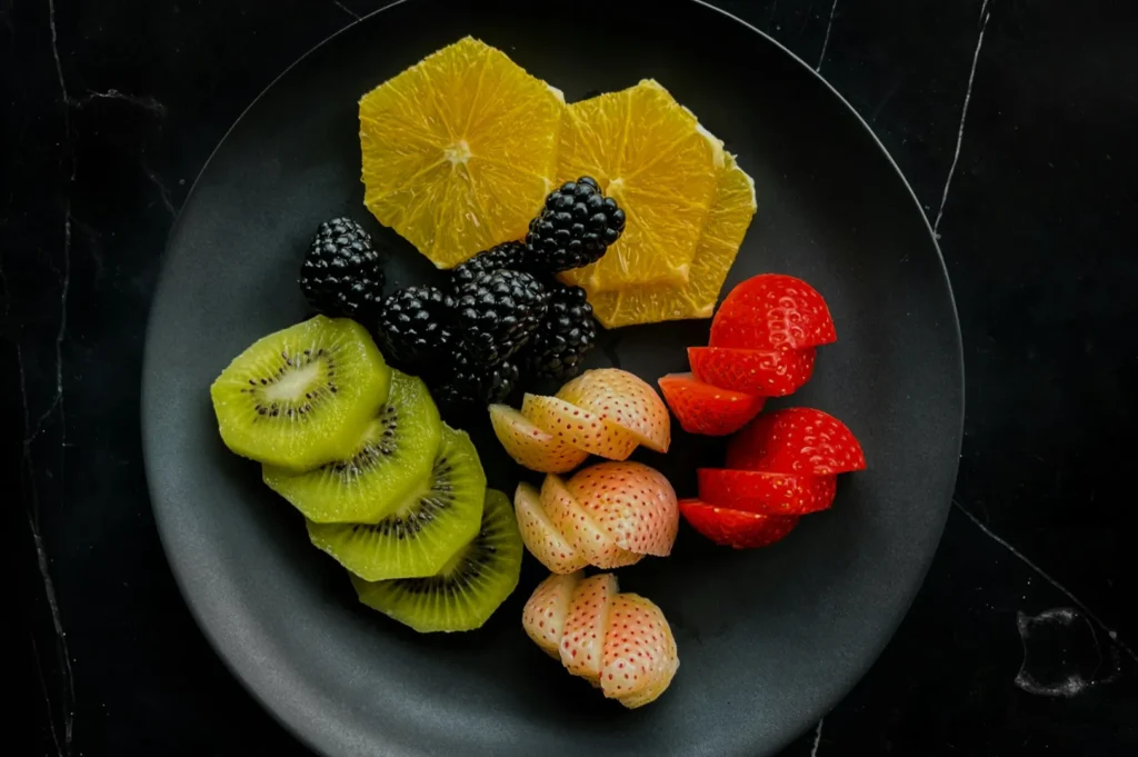 A black plate with sliced oranges, kiwi, blackberries, red and white strawberries arranged artistically, set on a dark marble background.