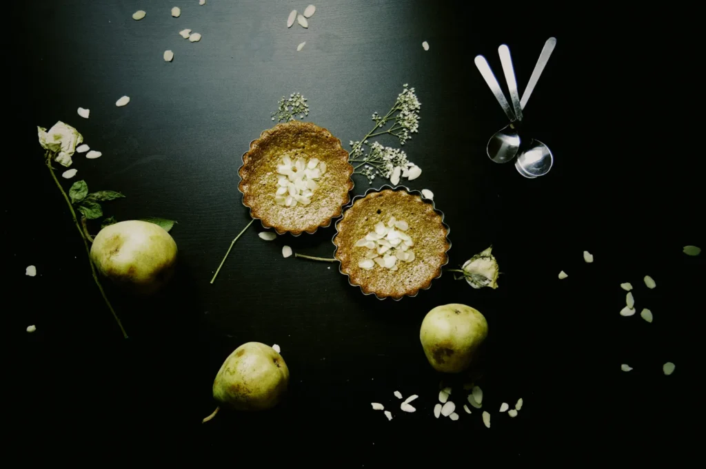Two rustic mini pies topped with almond slices sit on a dark surface, surrounded by three pears, white petals, and sprigs of flowers, creating an elegant, serene atmosphere.