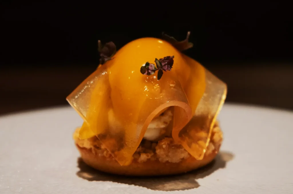A gourmet dessert featuring a round biscuit base topped with a glossy yellow dome and translucent sheet, adorned with tiny purple flowers on a white plate.