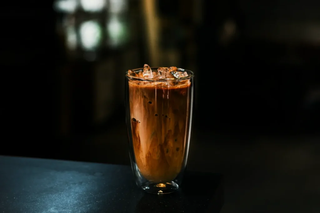 A glass of iced coffee with swirling milk sits on a dark countertop in soft focus lighting, creating a cozy, inviting atmosphere.