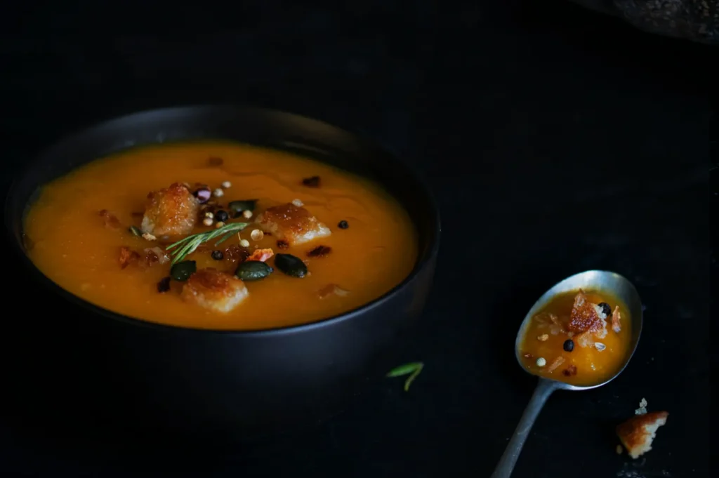 A dark bowl of creamy orange soup garnished with croutons, seeds, and herbs. Nearby, a spoon holds a small serving of the same soup. The mood is cozy and inviting.