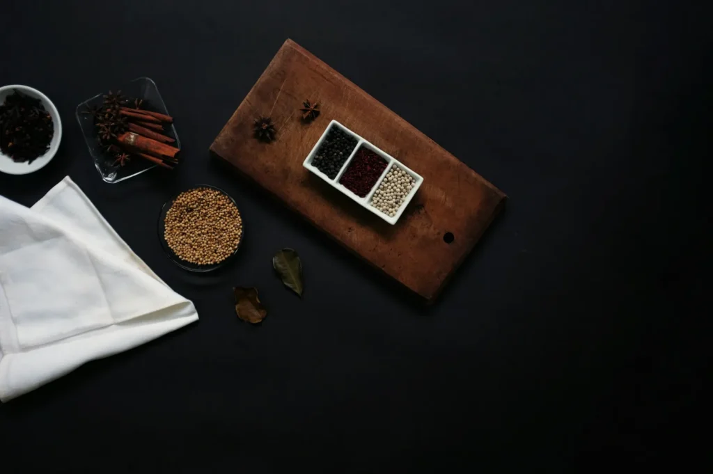 Dark, moody kitchen scene with spices. Wooden board holds three bowls of colorful spices. Nearby are whole spices, a white napkin, and bowls of seeds.
