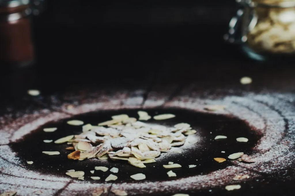 A pile of sliced almonds dusted with powdered sugar is on a dark wooden surface. The background is blurred, creating a warm, rustic atmosphere.