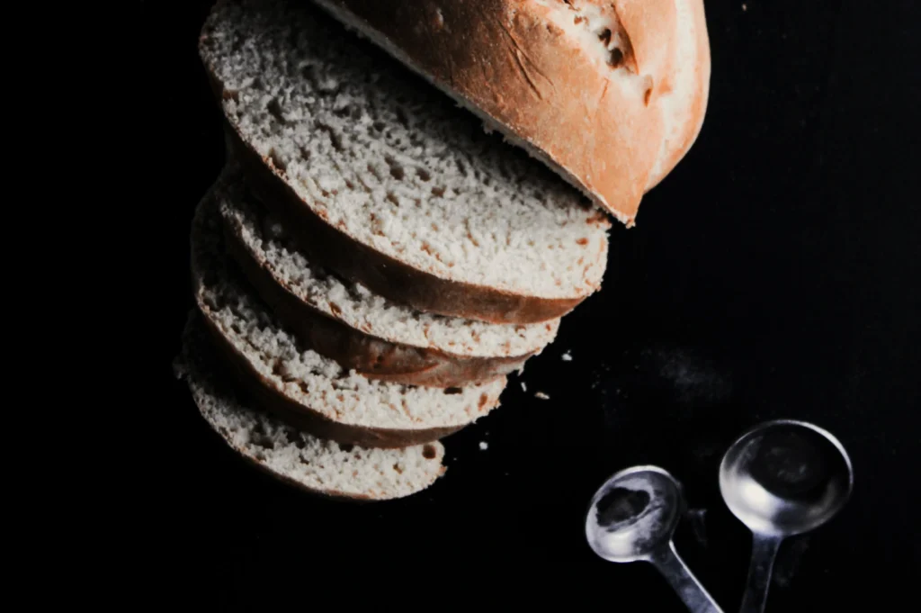 Sliced loaf of white bread on a dark surface, accompanied by two metallic measuring spoons. The scene evokes simplicity and home baking.