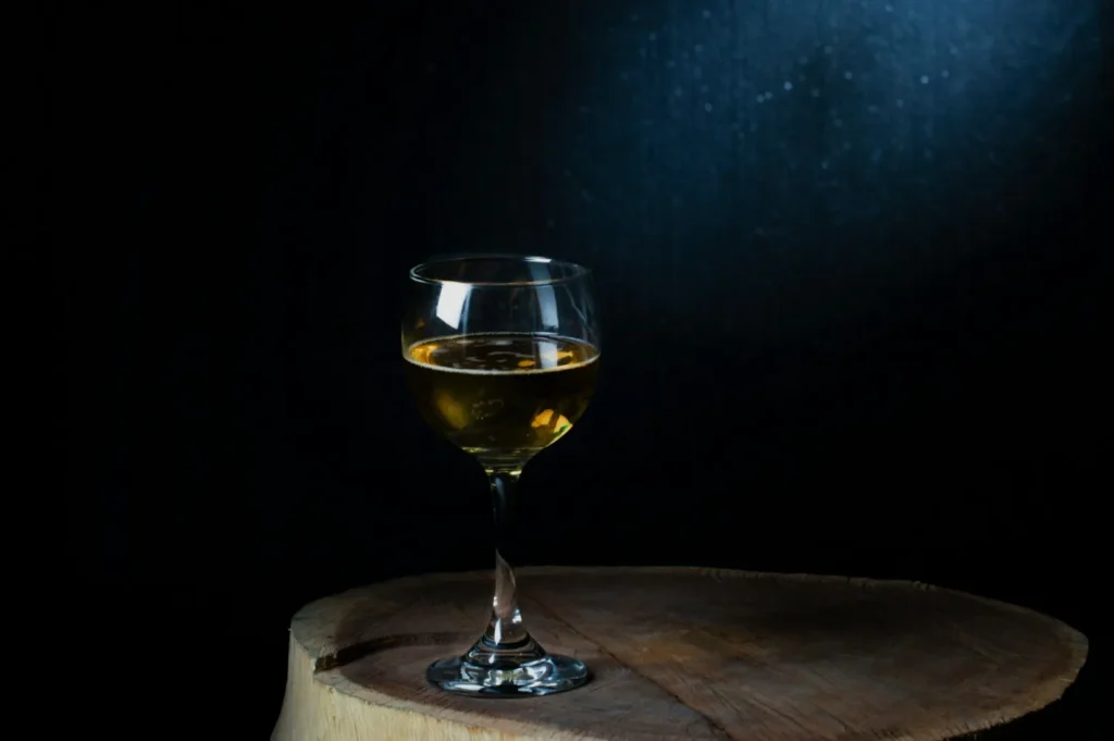 A wine glass filled with amber liquid sits on a wooden table. The dim lighting and dark background create an intimate and elegant atmosphere.