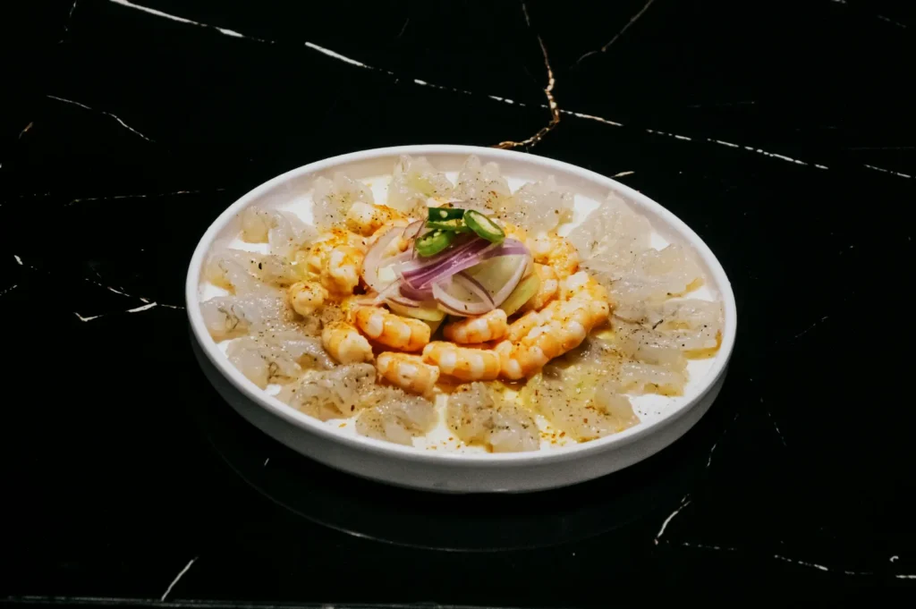 A white plate on a dark marbled surface displays a seafood dish of shrimp and thinly sliced white fish, garnished with onion and green peppers.