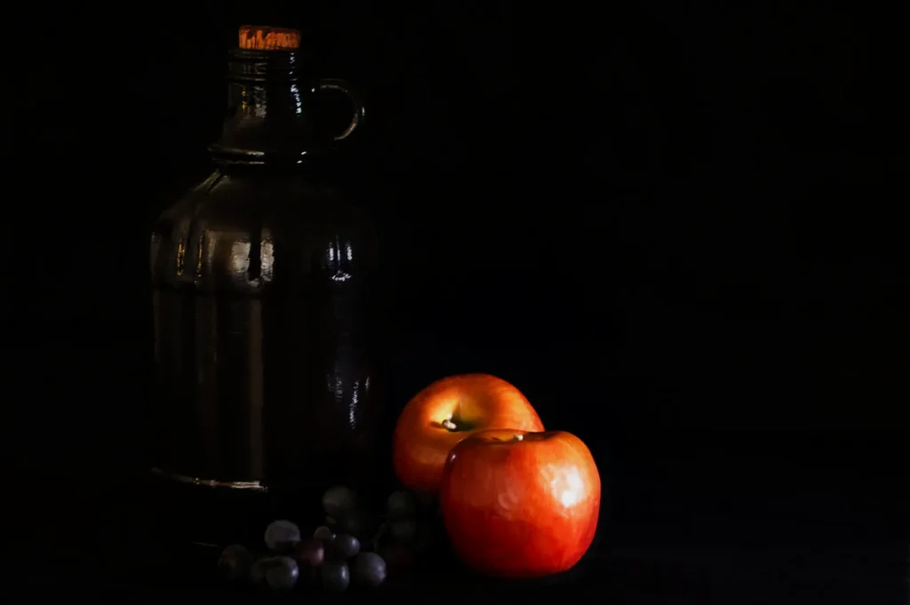 A dark glass jug with a cork sits beside two shiny red apples and a cluster of grapes against a black background, conveying a classic, moody still-life.