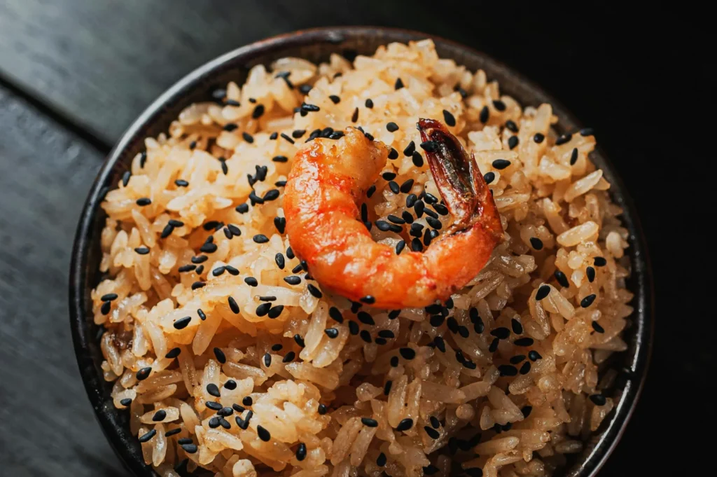 A bowl of glistening rice topped with a cooked shrimp and sprinkled with black sesame seeds, set on a dark wooden surface.