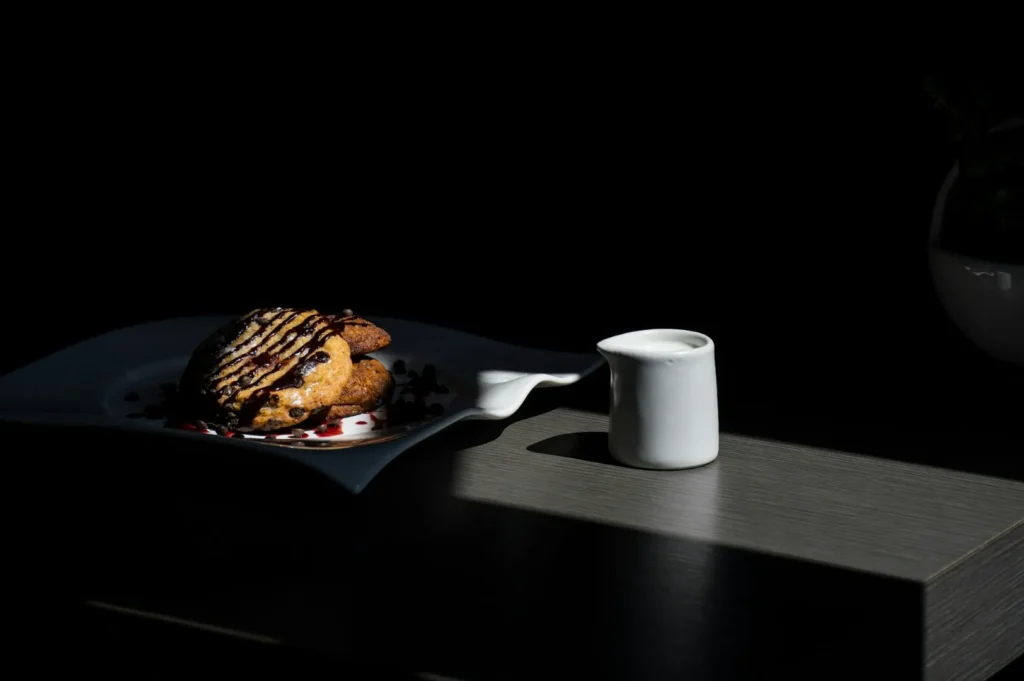 Cookies drizzled with chocolate sauce on a decorative plate are illuminated by soft light, with a small white jug beside them on a dark table.