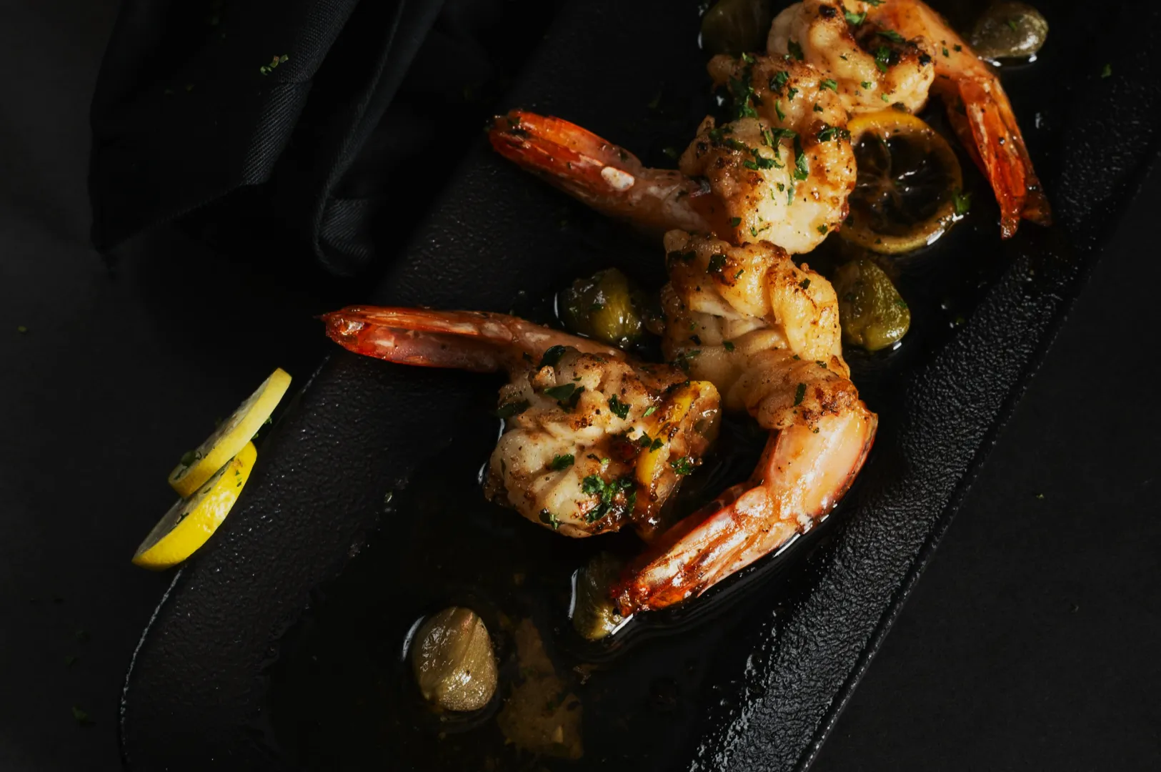 Grilled shrimp garnished with herbs, served on a black platter with lemon wedges and capers. The dish is presented on a dark, elegant background.