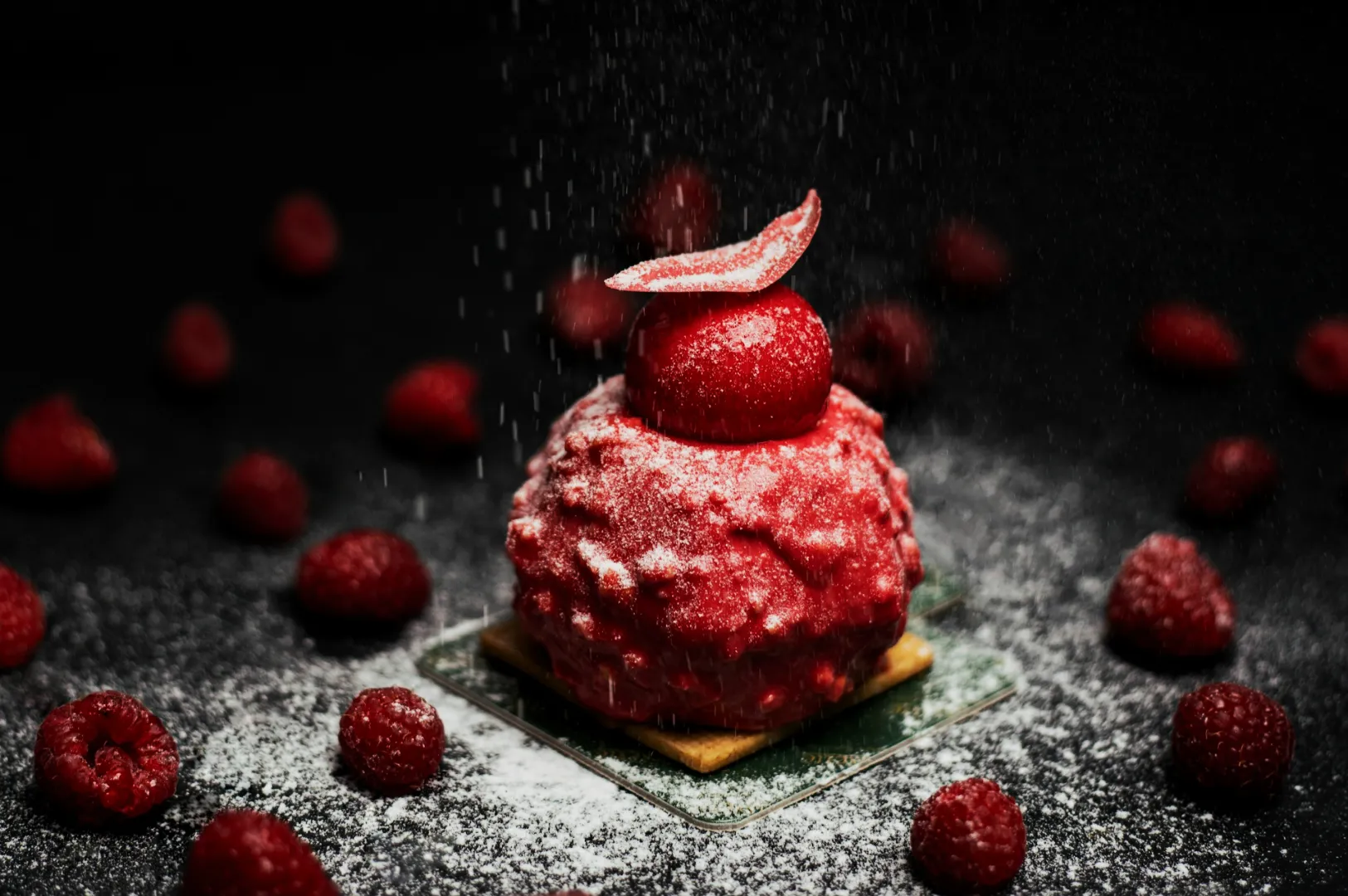 A vibrant red dessert topped with a raspberry and dusted with powdered sugar sits on a square base. Surrounded by scattered raspberries on a dark surface.