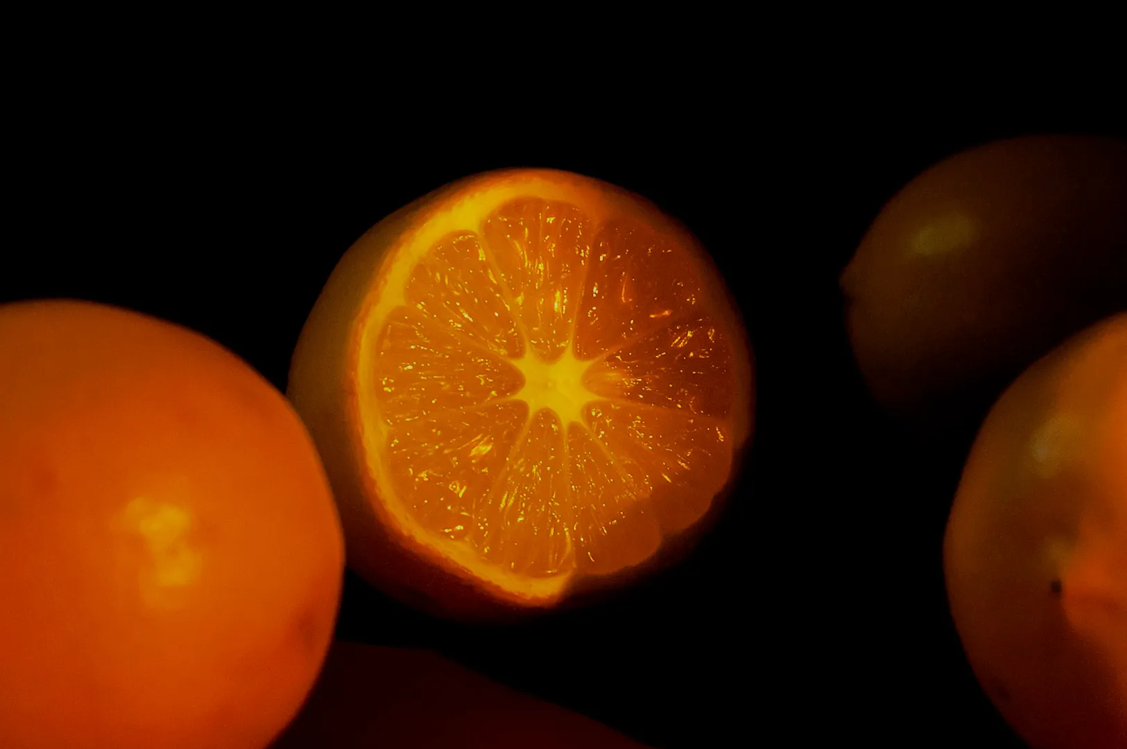 Close-up of a glowing orange sliced in half against a dark background, with whole oranges surrounding it. The lighting creates a warm, vibrant tone.
