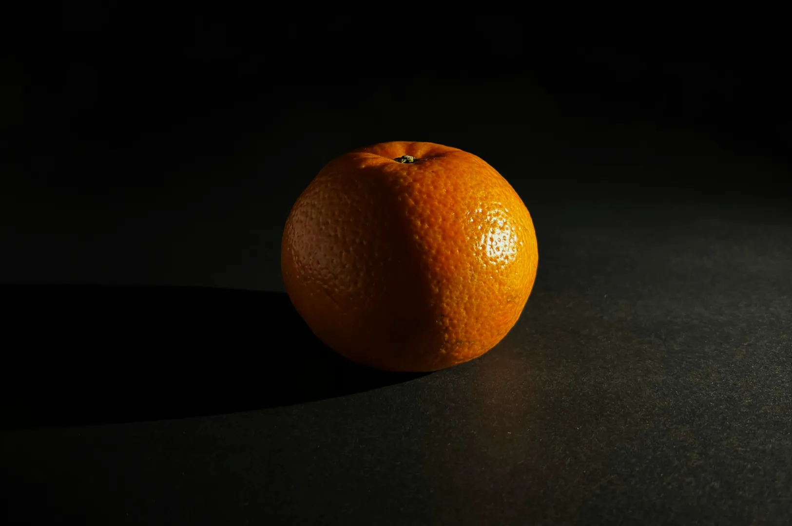 A single orange on a dark surface, highlighted by dramatic lighting from the left, casting a long shadow to the right, creating a minimalist and moody atmosphere.
