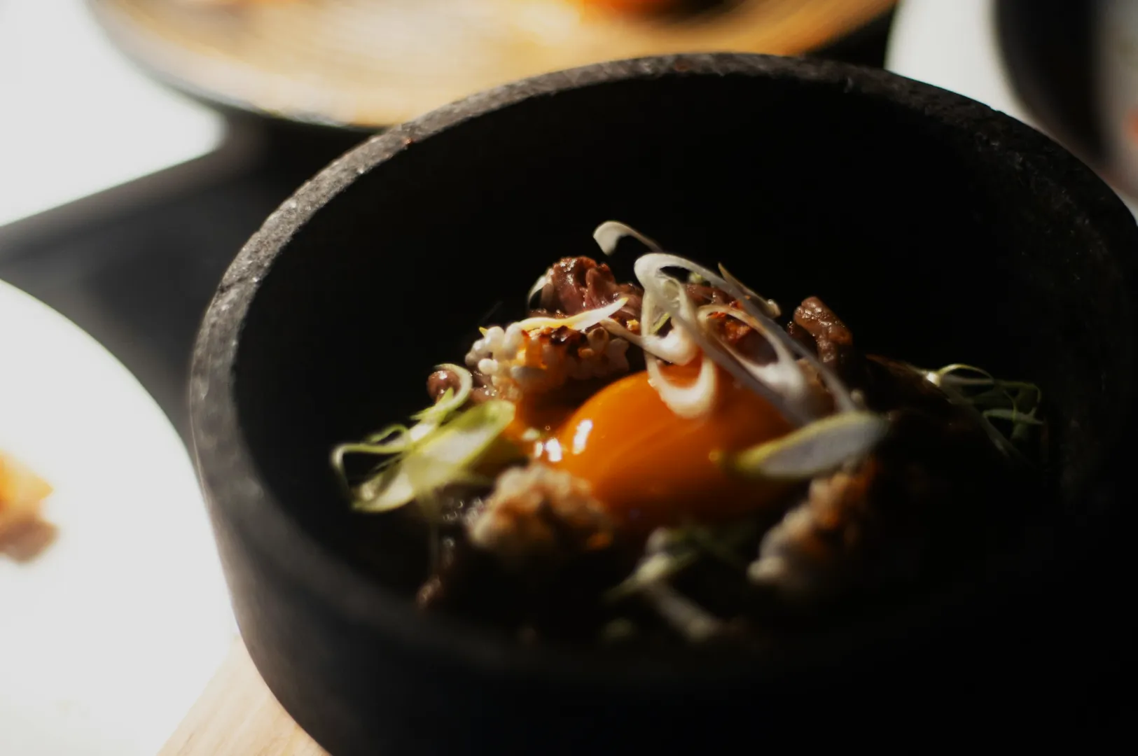 Close-up of a dish in a black bowl featuring a raw egg yolk, sliced green onions, and mixed ingredients, creating a warm, inviting atmosphere.