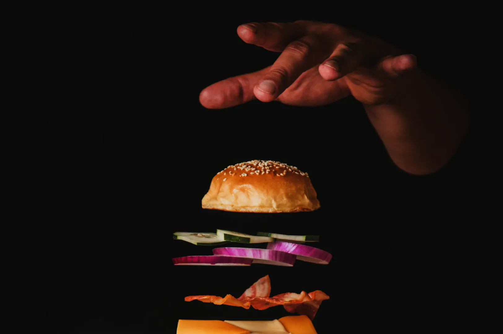 Floating burger ingredients, including a sesame bun, cucumbers, red onions, bacon, and cheese, against a dark background with a hand above.