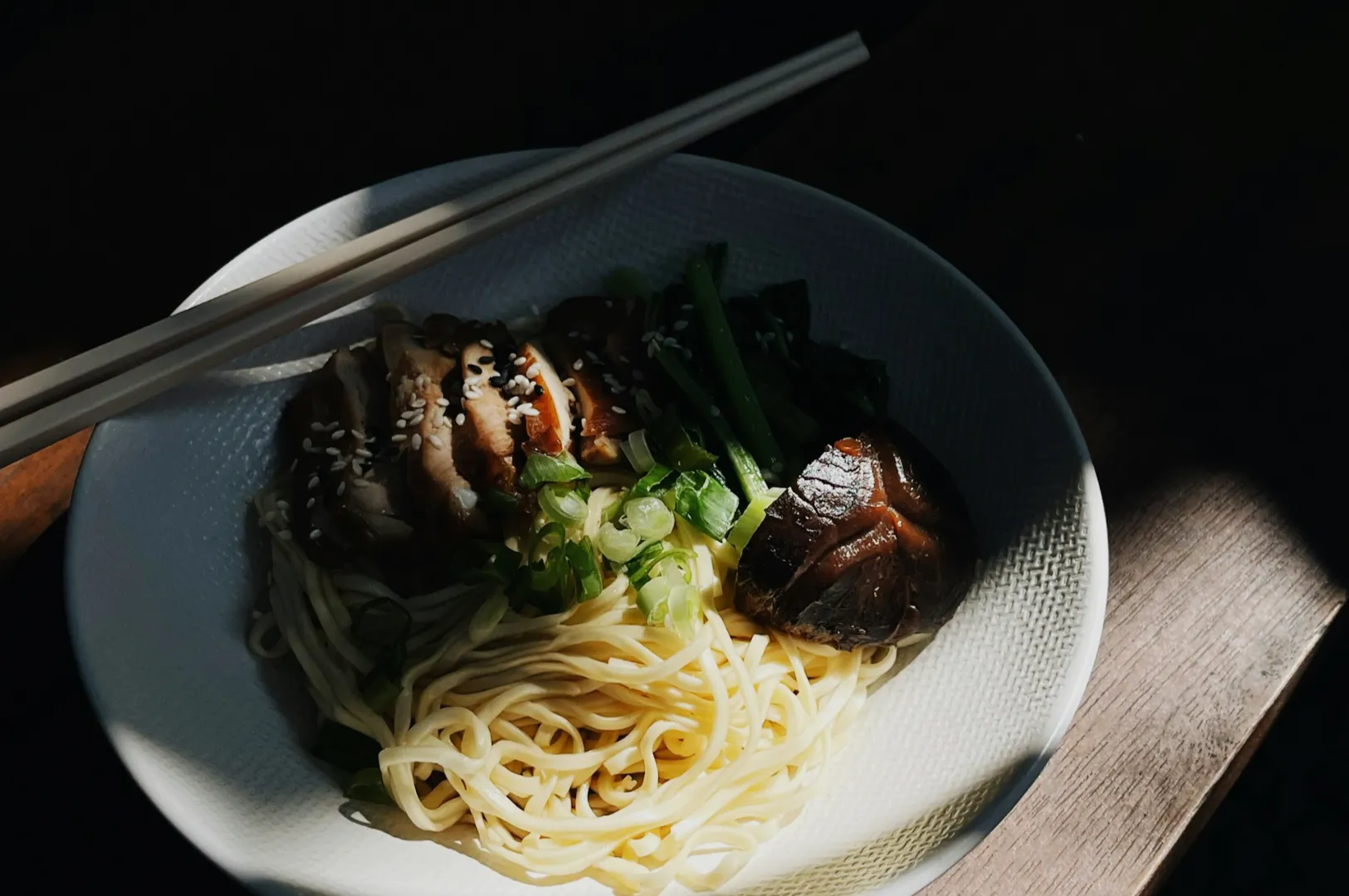 A white bowl of noodles topped with sliced chicken, mushrooms, and green vegetables. Chopsticks rest on the bowl, with sunlight casting shadows.