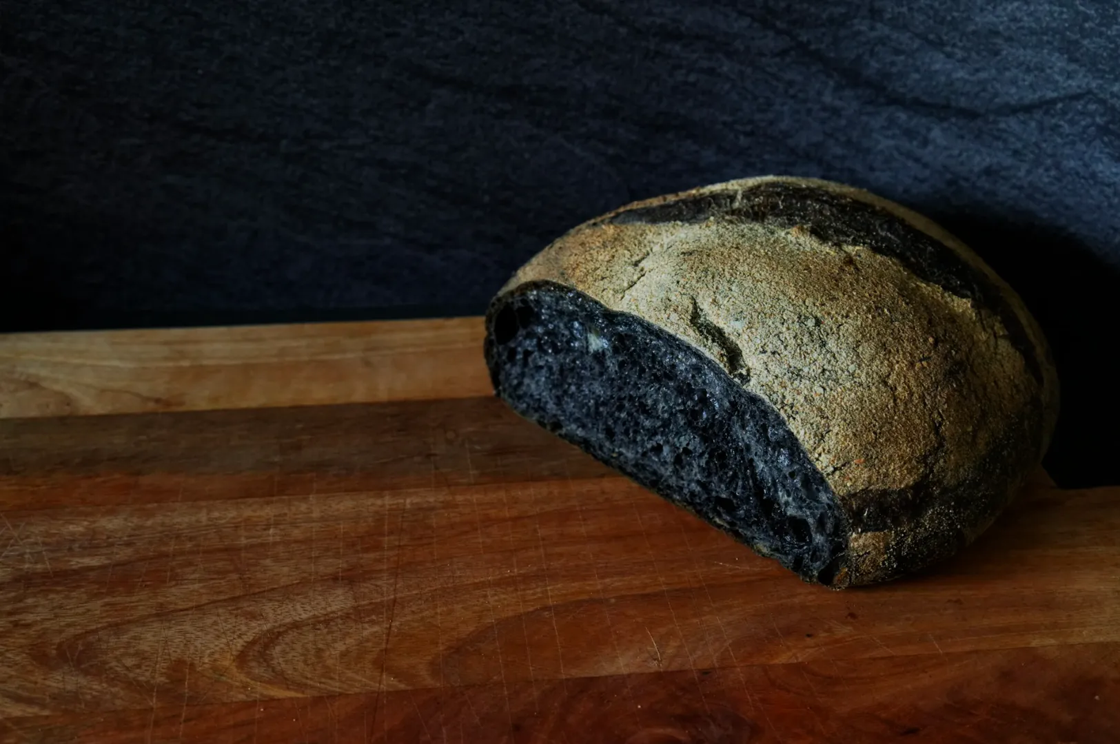 A rustic loaf of black charcoal bread with a cracked crust sits on a wooden cutting board against a dark background, conveying an artisanal, earthy vibe.