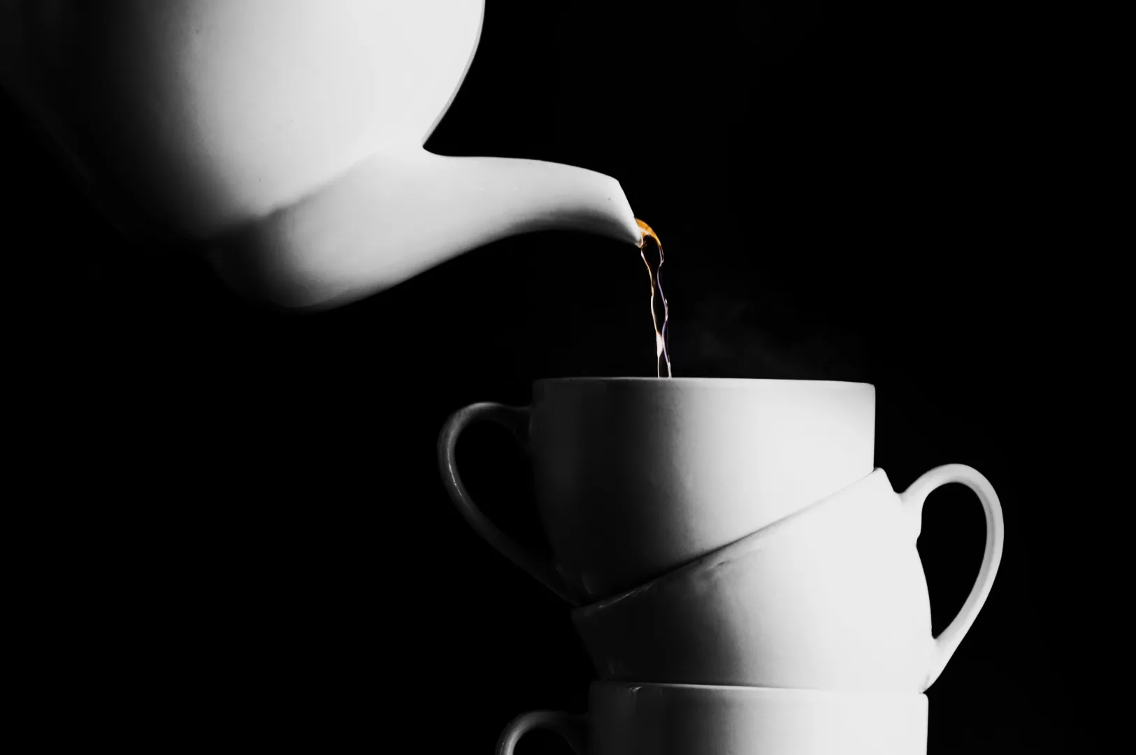 White teapot pouring steaming liquid into a stack of three white teacups against a black background, creating a minimalist and elegant mood.