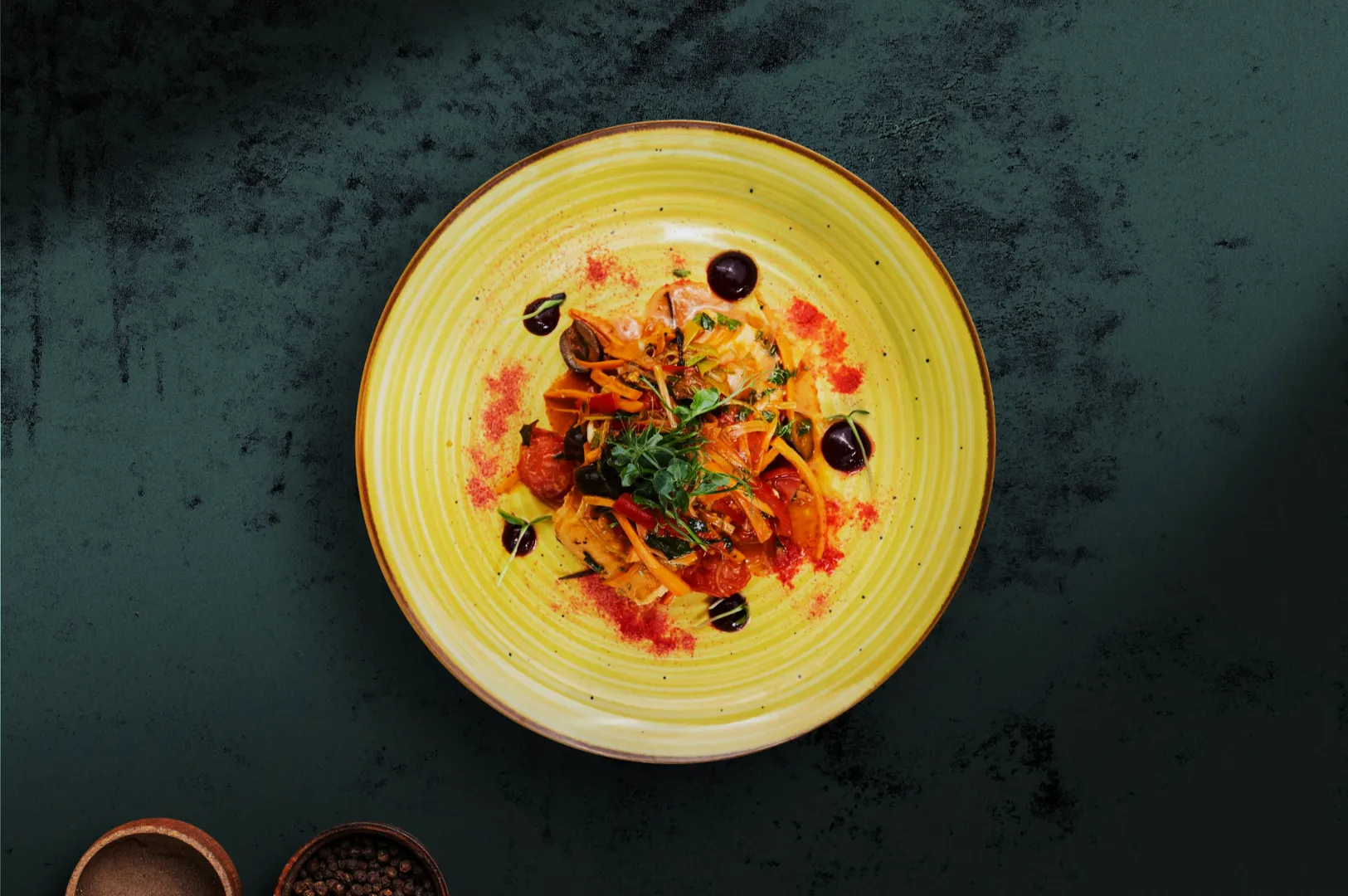 A colorful dish on a yellow plate with greens, carrots, and dark sauce dots, garnished with herbs. The setting is on a textured, dark surface.