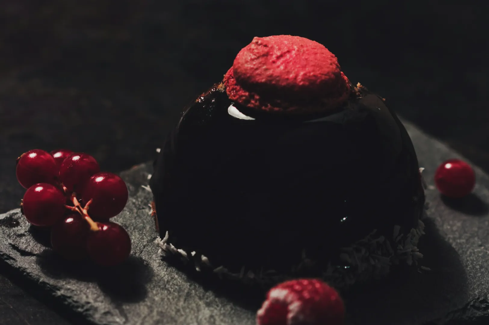 Elegant dessert on dark slate, featuring a glossy chocolate dome topped with a red macaron. Cluster of red currants adds a pop of color.