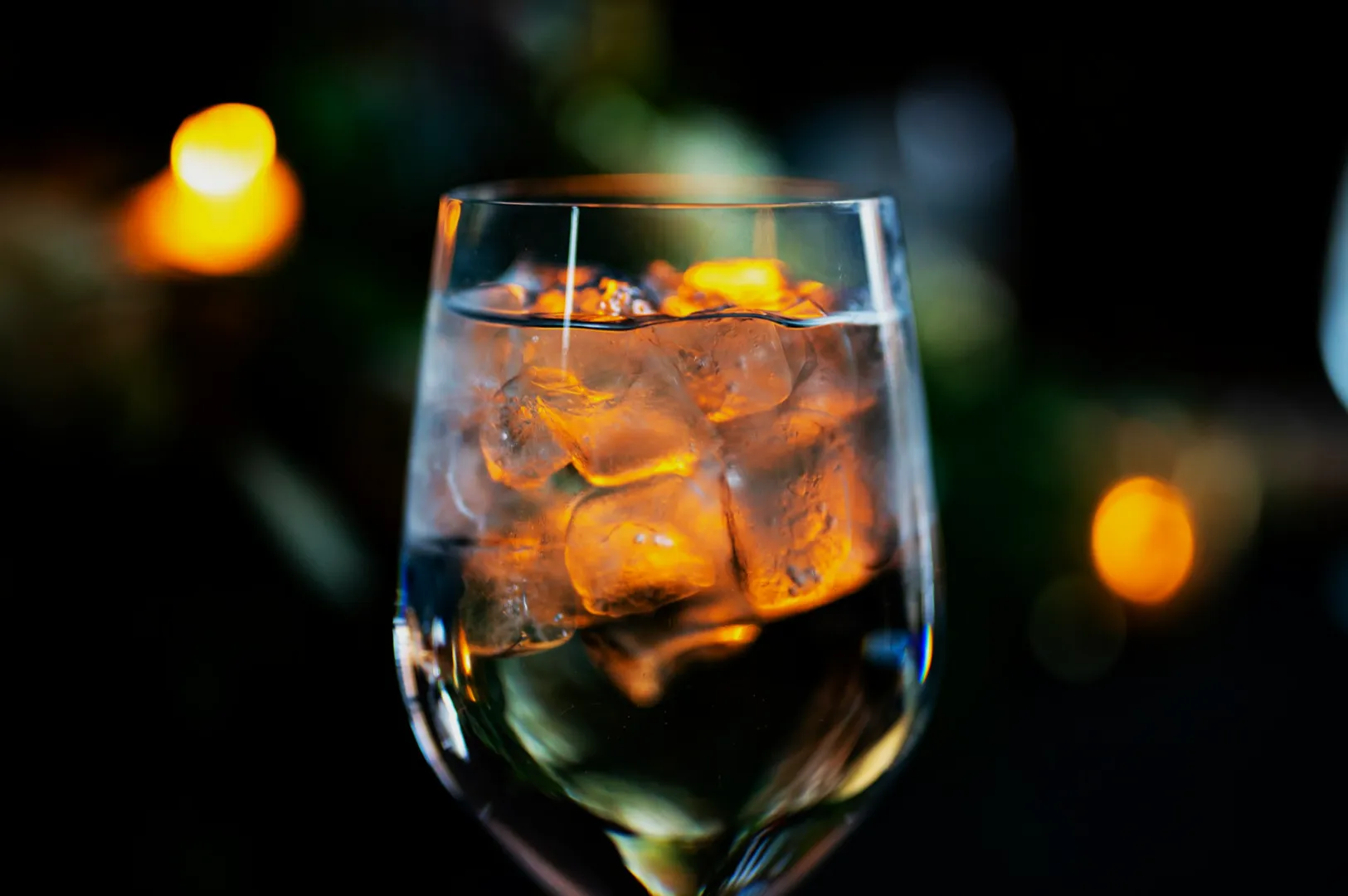 A wine glass filled with ice cubes reflects warm, yellow lights, set against a dark, blurred background with a cozy and elegant ambiance.