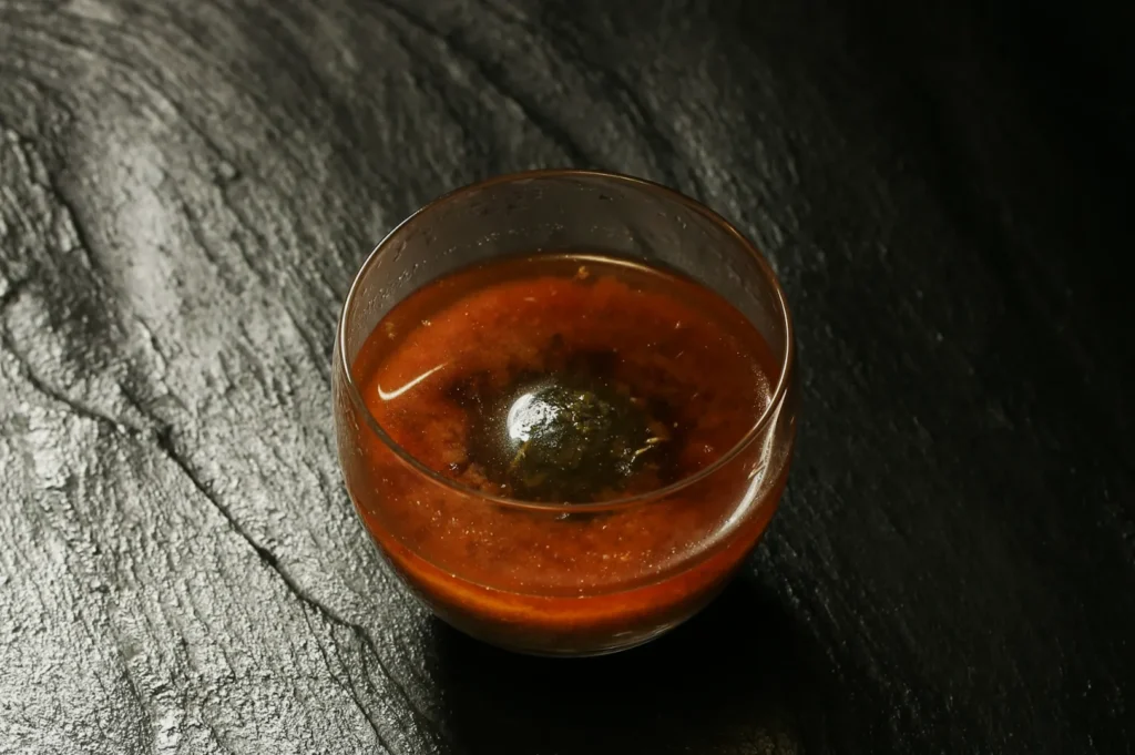 A glass of brown liquid with a floating green-black sphere, set against a textured black stone surface, creating a mysterious and elegant vibe.